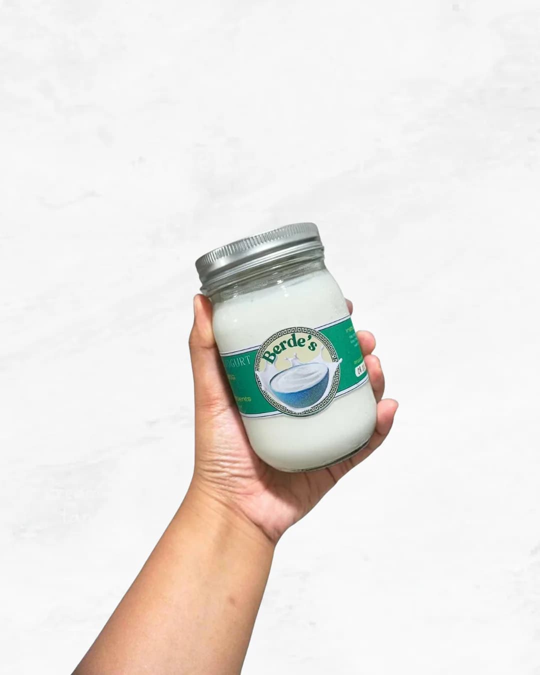 A hand holding a jar of Berde's plain Greek yogurt, thick and white through the glass.