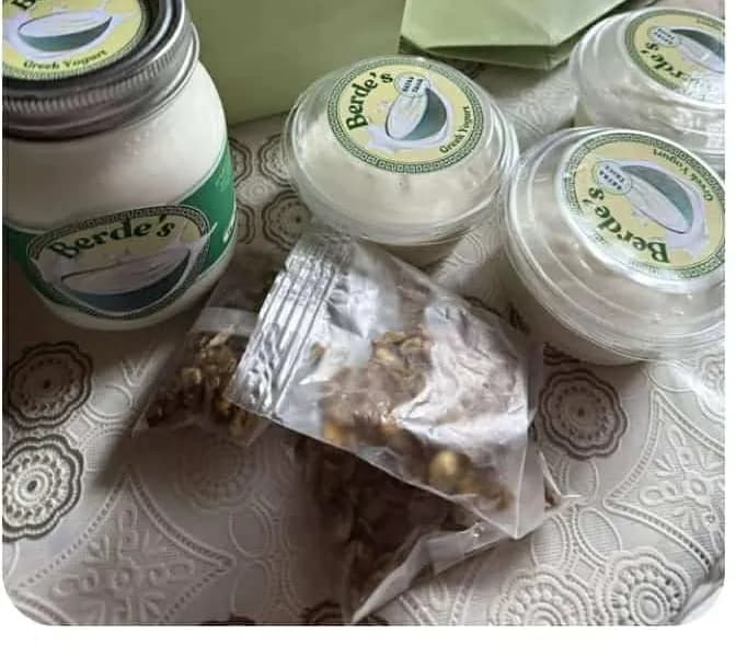 A Berde's jar alongside several sealed cups of yogurt and a bag of granola on a lace tablecloth.