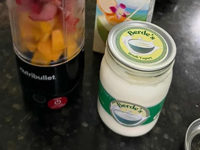 A jar of Berde's plain yogurt on a kitchen counter beside a blender cup filled with chopped mango and berries.